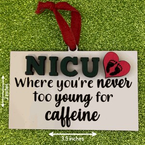 K&ouml;nnte beinhalten: Ein wei&szlig;er Holz-Anh&auml;nger mit dem Text "NICU Where you're never too young for caffeine" in schwarzer Schrift. Ein pinkfarbenes Herz mit schwarzen Baby-Fu&szlig;abdr&uuml;cken ist oben rechts angebracht. Der Anh&auml;nger ist 8,9 cm breit und 5,1 cm hoch.
