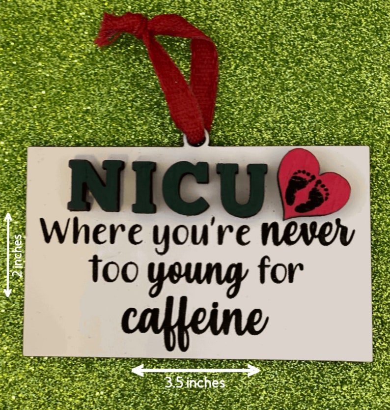 K&ouml;nnte beinhalten: Ein wei&szlig;er Holz-Anh&auml;nger mit dem Text "NICU Where you're never too young for caffeine" in schwarzer Schrift. Ein pinkfarbenes Herz mit schwarzen Baby-Fu&szlig;abdr&uuml;cken ist oben rechts angebracht. Der Anh&auml;nger ist 8,9 cm breit und 5,1 cm hoch.