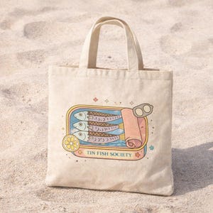 Mediterranean coastal sardine tote bag standing on beach sand with vintage tin fish illustration