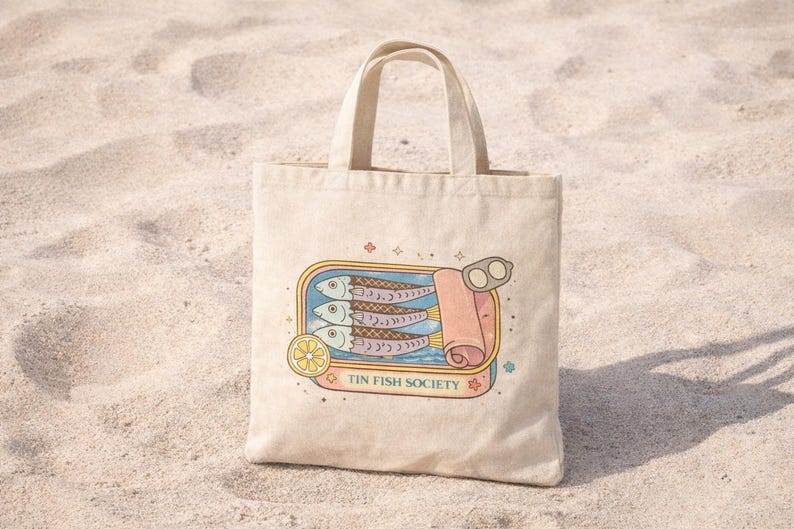 Mediterranean coastal sardine tote bag standing on beach sand with vintage tin fish illustration