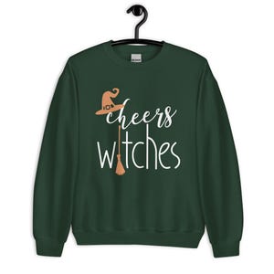 Op de afbeelding: Een donker bosgroen sweatshirt met lange mouwen en een grafische print in wit en lichtoranje. De tekst luidt cheers witches, met een lichtoranje heksenhoed boven het woord cheers en een lichtoranje bezemsteel die de letter i vervangt.