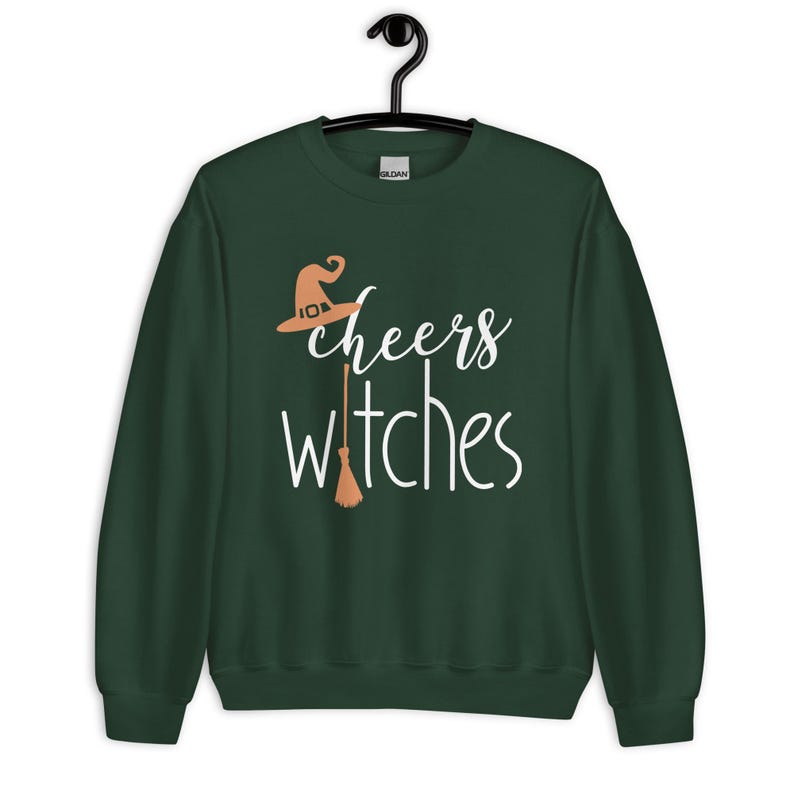 Op de afbeelding: Een donker bosgroen sweatshirt met lange mouwen en een grafische print in wit en lichtoranje. De tekst luidt cheers witches, met een lichtoranje heksenhoed boven het woord cheers en een lichtoranje bezemsteel die de letter i vervangt.