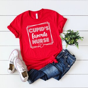 May include: Red t-shirt with white text that says "Cupid's Favorite Nurse" with a heart and arrow graphic.