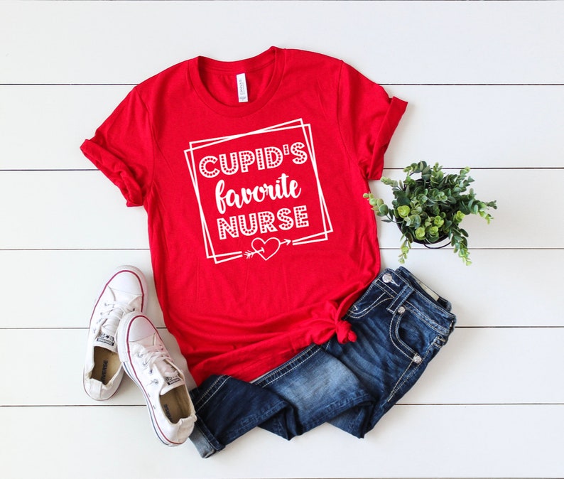 May include: Red t-shirt with white text that says "Cupid's Favorite Nurse" with a heart and arrow graphic.