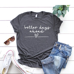 May include: A dark grey t-shirt with the text "better days ahead" in white lettering with a small heart underneath. The shirt is laid out on a white wooden surface with a pair of sunglasses, a pair of white trainers, and a pair of blue jeans.