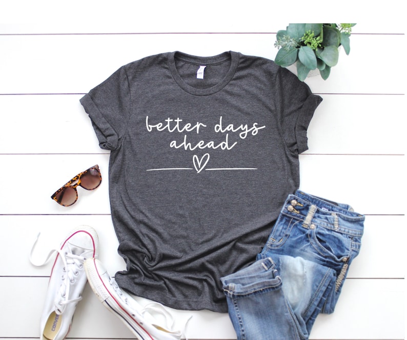 May include: A dark grey t-shirt with the text "better days ahead" in white lettering with a small heart underneath. The shirt is laid out on a white wooden surface with a pair of sunglasses, a pair of white trainers, and a pair of blue jeans.