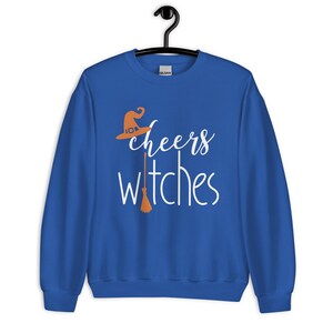 Op de afbeelding: Een koningsblauw sweatshirt met lange mouwen en een witte grafische opdruk met de tekst cheers witches. Het ontwerp bevat een oranje heksenhoed boven het woord cheers en een oranje bezemsteel die de letter t in witches vervangt.