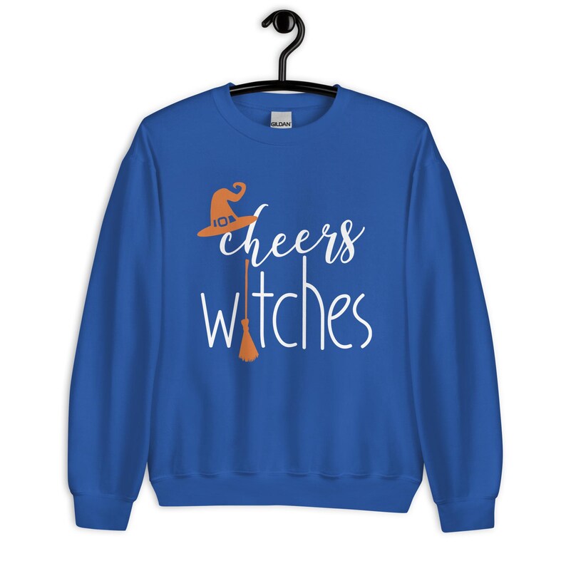 Op de afbeelding: Een koningsblauw sweatshirt met lange mouwen en een witte grafische opdruk met de tekst cheers witches. Het ontwerp bevat een oranje heksenhoed boven het woord cheers en een oranje bezemsteel die de letter t in witches vervangt.