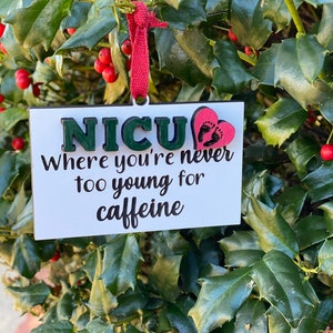 K&ouml;nnte beinhalten: Ein wei&szlig;er Holzschmuck mit dem Text "NICU Where you're never too young for caffeine" in schwarzer Schrift. Ein rotes Herz mit schwarzen Fu&szlig;abdr&uuml;cken ist an dem Schmuck befestigt.