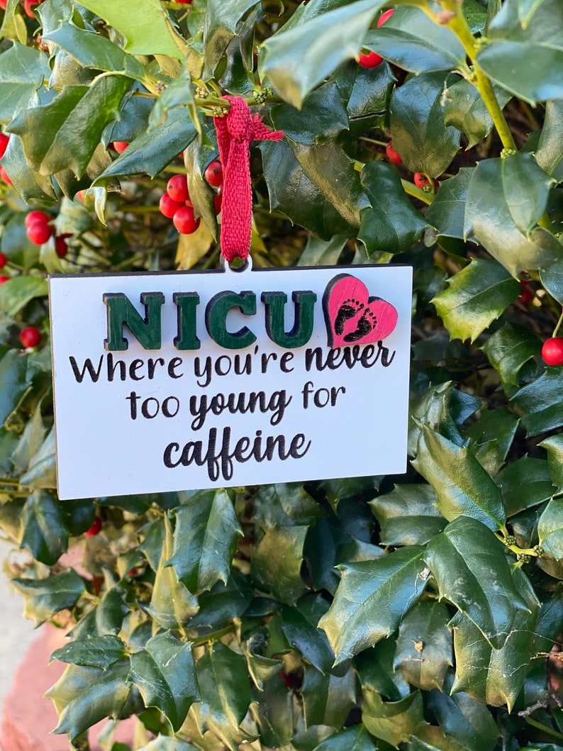 K&ouml;nnte beinhalten: Ein wei&szlig;er Holzschmuck mit dem Text "NICU Where you're never too young for caffeine" in schwarzer Schrift. Ein rotes Herz mit schwarzen Fu&szlig;abdr&uuml;cken ist an dem Schmuck befestigt.