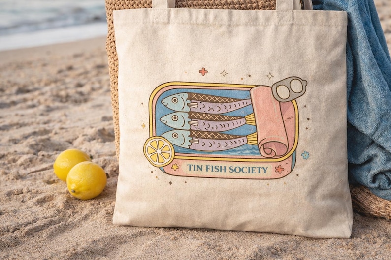 Sardine tote bag with Tin Fish Society design on beach sand with lemons and coastal aesthetic styling