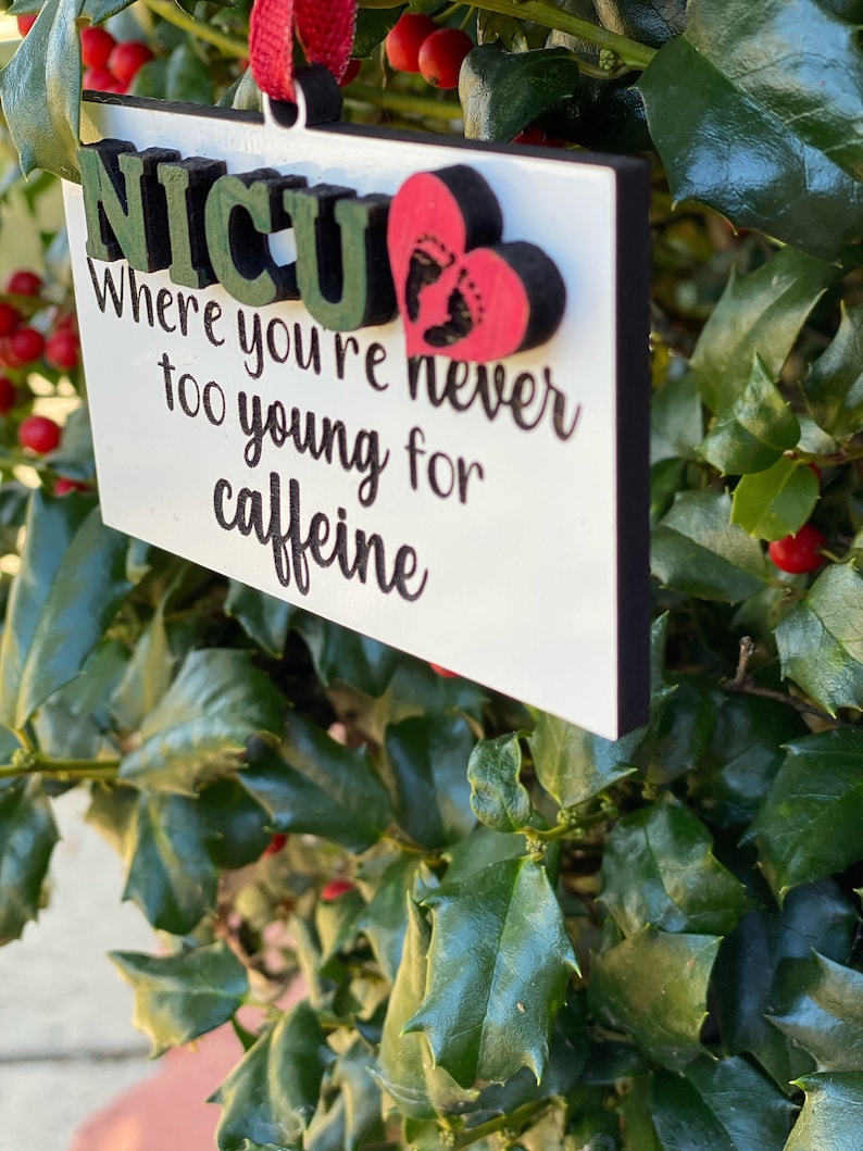 K&ouml;nnte beinhalten: Wei&szlig;er Holzanh&auml;nger mit dem Text "NICU Where you're never too young for caffeine" und einem roten Herz mit einem Fu&szlig;abdruck-Design.