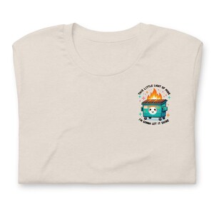 Puede incluir: Una camiseta beige claro con un gr&aacute;fico de un contenedor de basura verde en llamas con el texto "This little light of mine, I'm gonna let it shine".