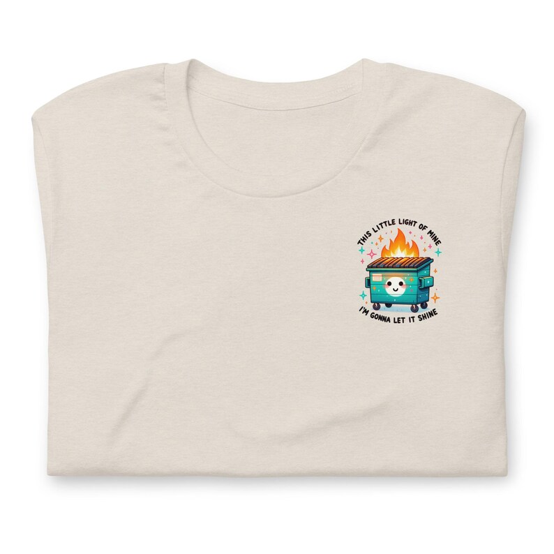 Puede incluir: Una camiseta beige claro con un gr&aacute;fico de un contenedor de basura verde en llamas con el texto "This little light of mine, I'm gonna let it shine".