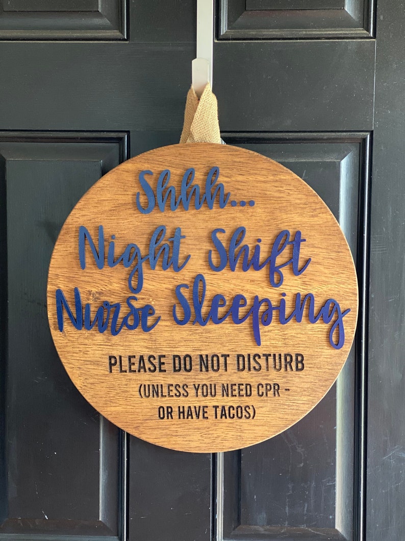 May include: A round wooden sign with the text "Shhh... Night Shift Nurse Sleeping" in blue lettering. The sign also includes the text "PLEASE DO NOT DISTURB (UNLESS YOU NEED CPR - OR HAVE TACOS)" in black lettering.