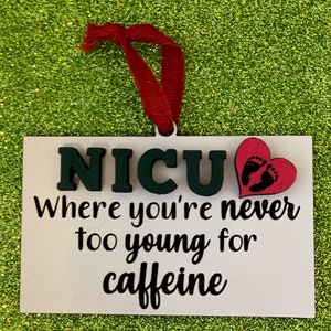 K&ouml;nnte beinhalten: Wei&szlig;er Holzanh&auml;nger mit dem Text "NICU Where you're never too young for caffeine" und einem rosa Herz mit Fu&szlig;abdr&uuml;cken.