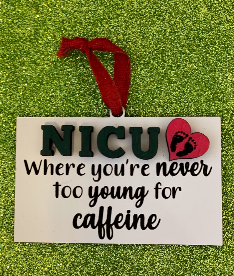 K&ouml;nnte beinhalten: Wei&szlig;er Holzanh&auml;nger mit dem Text "NICU Where you're never too young for caffeine" und einem rosa Herz mit Fu&szlig;abdr&uuml;cken.