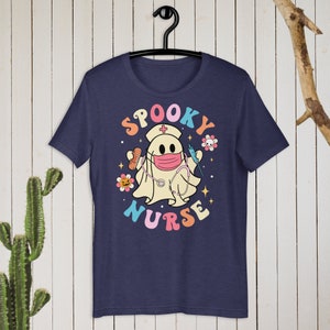 以下が含まれることがあります： ネイビーブルーのTシャツ。看護師の格好をした幽霊の漫画が描かれています。幽霊はマスク、帽子、聴診器を着用し、医療用品を持っています。「Spooky Nurse」の文字はカラフルなレトロスタイルです。
