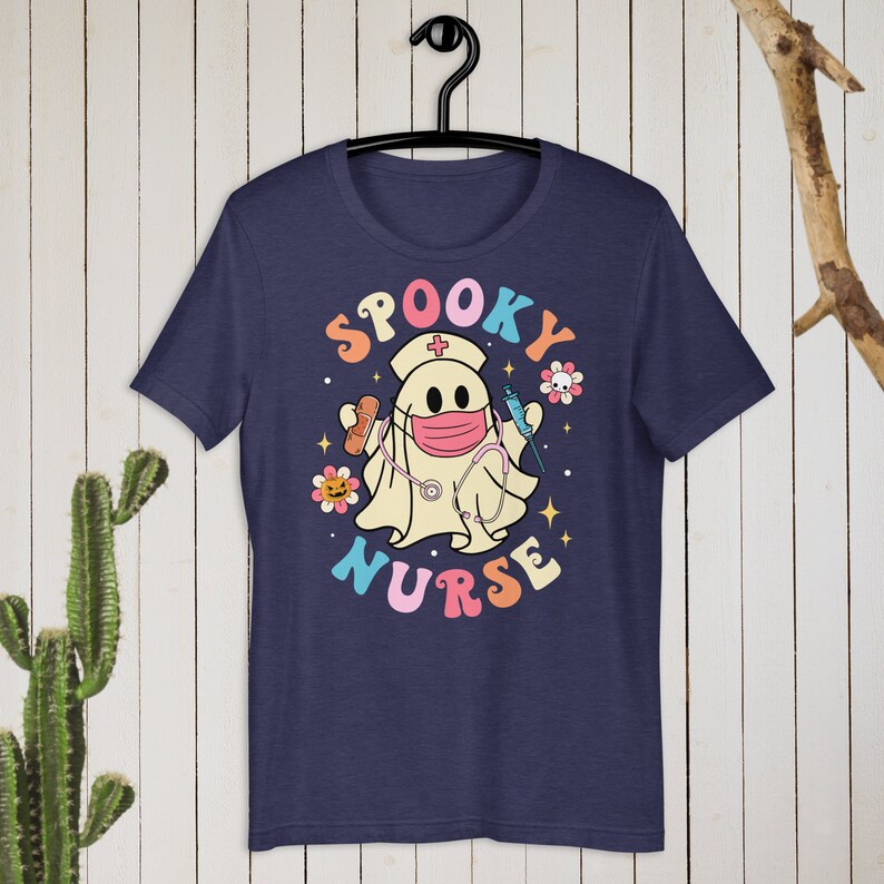 以下が含まれることがあります： ネイビーブルーのTシャツ。看護師の格好をした幽霊の漫画が描かれています。幽霊はマスク、帽子、聴診器を着用し、医療用品を持っています。「Spooky Nurse」の文字はカラフルなレトロスタイルです。