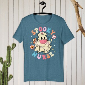以下が含まれることがあります： 水色のTシャツ。看護師の格好をした漫画の幽霊が描かれています。幽霊はマスクをして、包帯と注射器を持っています。「SPOOKY NURSE」の文字がレトロなスタイルでカラフルに書かれています。Tシャツは黒いハンガーにかかっています。
