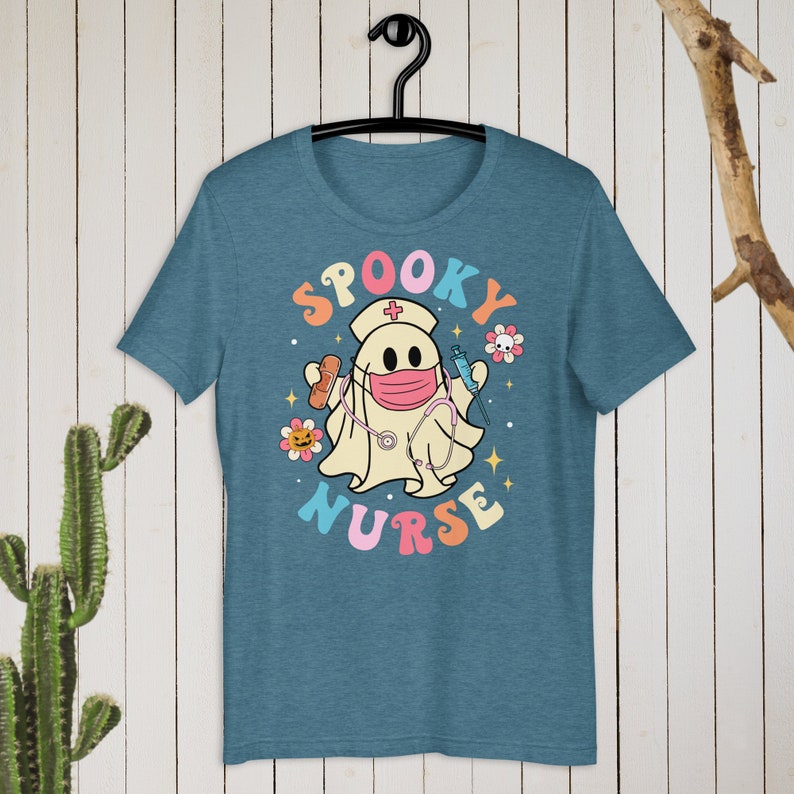 以下が含まれることがあります： 水色のTシャツ。看護師の格好をした漫画の幽霊が描かれています。幽霊はマスクをして、包帯と注射器を持っています。「SPOOKY NURSE」の文字がレトロなスタイルでカラフルに書かれています。Tシャツは黒いハンガーにかかっています。