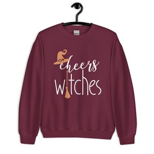 Op de afbeelding: Een bordeauxrood sweatshirt met lange mouwen en de witte tekst "cheers witches" op de voorzijde. Het ontwerp bevat een lichtoranje heksenhoed boven het woord "cheers" en een lichtoranje bezemsteel die de letter "t" in "witches" vervangt.