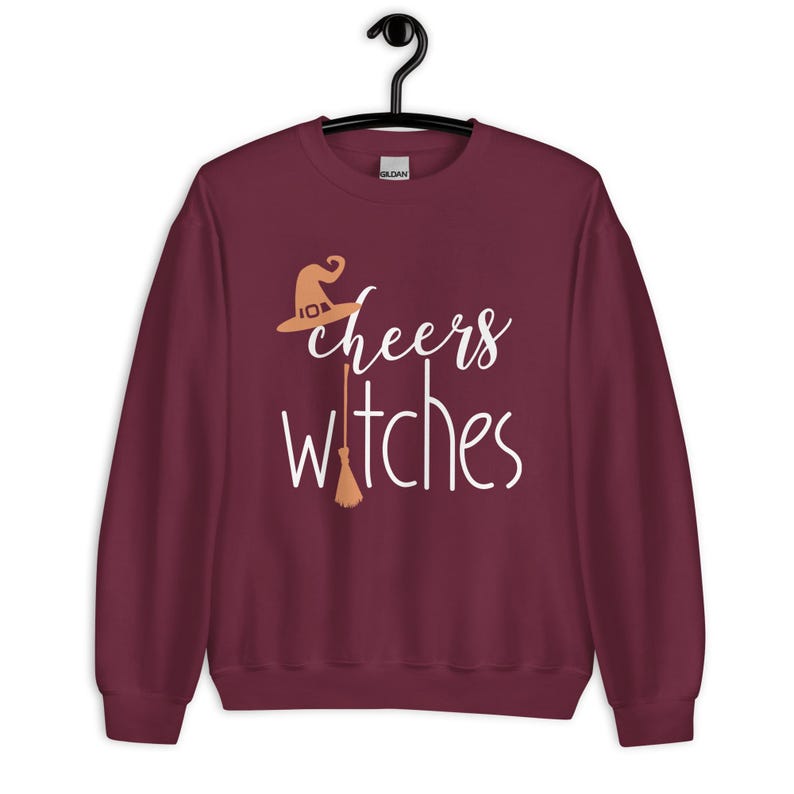 Op de afbeelding: Een bordeauxrood sweatshirt met lange mouwen en de witte tekst "cheers witches" op de voorzijde. Het ontwerp bevat een lichtoranje heksenhoed boven het woord "cheers" en een lichtoranje bezemsteel die de letter "t" in "witches" vervangt.