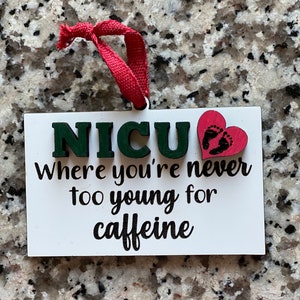 K&ouml;nnte beinhalten: Wei&szlig;er Holzanh&auml;nger mit dem Text "NICU Where you're never too young for caffeine" in schwarzer Schrift. Ein rotes Herz mit schwarzen Fu&szlig;abdr&uuml;cken ist an dem Anh&auml;nger befestigt.