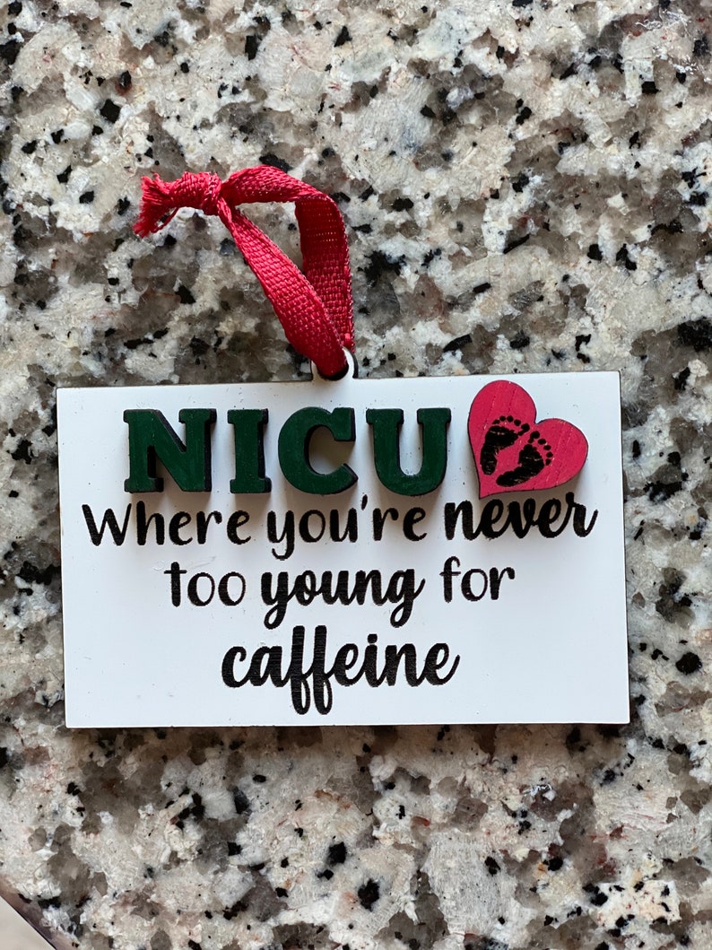 K&ouml;nnte beinhalten: Wei&szlig;er Holzanh&auml;nger mit dem Text "NICU Where you're never too young for caffeine" in schwarzer Schrift. Ein rotes Herz mit schwarzen Fu&szlig;abdr&uuml;cken ist an dem Anh&auml;nger befestigt.