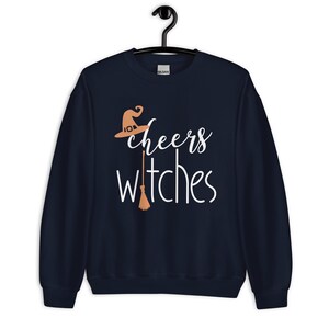 Op de afbeelding: Een marineblauw sweatshirt met lange mouwen en de witte tekst "cheers witches" op de voorzijde. Het ontwerp bevat een lichtoranje heksenhoed boven het woord cheers en een lichtoranje bezemsteel die de letter t vervangt.