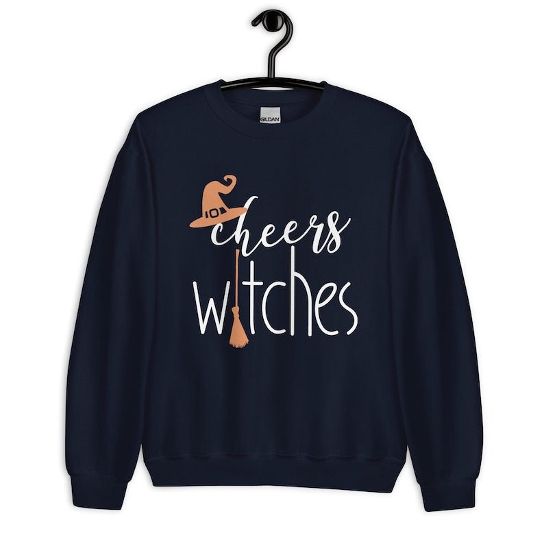 Op de afbeelding: Een marineblauw sweatshirt met lange mouwen en de witte tekst "cheers witches" op de voorzijde. Het ontwerp bevat een lichtoranje heksenhoed boven het woord cheers en een lichtoranje bezemsteel die de letter t vervangt.