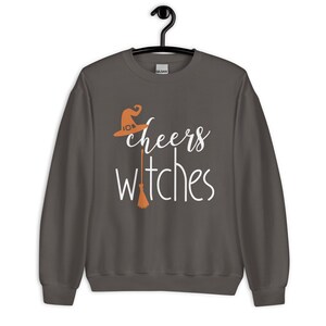 Op de afbeelding: Een donkergrijs sweatshirt met lange mouwen en de witte tekst "cheers witches" op de voorkant. Het ontwerp bevat een oranje heksenhoed boven het woord cheers en een oranje bezemsteel die de letter t in witches vervangt.
