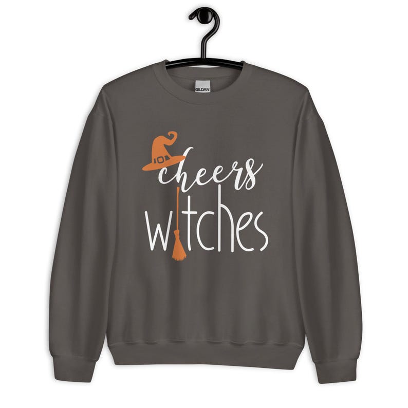 Op de afbeelding: Een donkergrijs sweatshirt met lange mouwen en de witte tekst "cheers witches" op de voorkant. Het ontwerp bevat een oranje heksenhoed boven het woord cheers en een oranje bezemsteel die de letter t in witches vervangt.