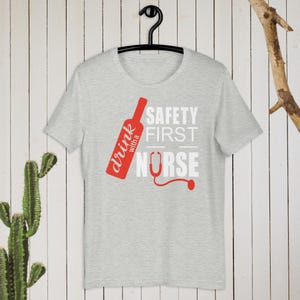 K&ouml;nnte beinhalten: Ein graues T-Shirt mit einer roten Weinflaschengrafik und dem Text "drink with a nurse" und "safety first" in Wei&szlig;.