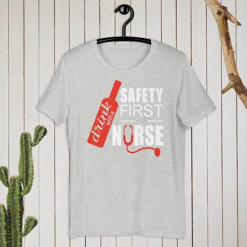 K&ouml;nnte beinhalten: Ein graues T-Shirt mit einer roten Weinflaschengrafik und dem Text "drink with a nurse" und "safety first" in Wei&szlig;.