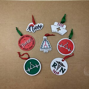 K&ouml;nnte beinhalten: Sechs Weihnachtsbaumschmuck mit einem Krankenschwester-Motiv. Der Schmuck ist rund und aus Holz. Er zeigt verschiedene Designs, darunter einen Weihnachtsbaum, ein Herz mit einer Herzschlaglinie und die Worte "Krankenschwester" und "RN".