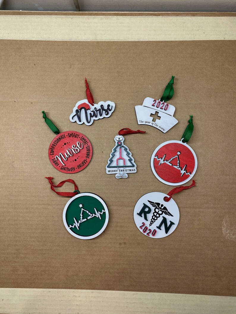 K&ouml;nnte beinhalten: Sechs Weihnachtsbaumschmuck mit einem Krankenschwester-Motiv. Der Schmuck ist rund und aus Holz. Er zeigt verschiedene Designs, darunter einen Weihnachtsbaum, ein Herz mit einer Herzschlaglinie und die Worte "Krankenschwester" und "RN".