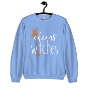 Op de afbeelding: Een lichtblauw sweatshirt met ronde hals en de witte tekst cheers witches. Het ontwerp bevat een oranje heksenhoed boven het woord cheers en een oranje bezemsteel die de letter i in het woord witches vervangt.