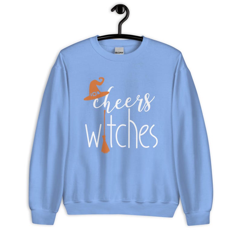 Op de afbeelding: Een lichtblauw sweatshirt met ronde hals en de witte tekst cheers witches. Het ontwerp bevat een oranje heksenhoed boven het woord cheers en een oranje bezemsteel die de letter i in het woord witches vervangt.
