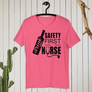 K&ouml;nnte beinhalten: Pinkes T-Shirt mit einem schwarzen Grafikdesign, auf dem "drink with a nurse" und "safety first" mit einem Stethoskop-Motiv steht.