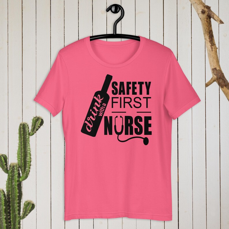 K&ouml;nnte beinhalten: Pinkes T-Shirt mit einem schwarzen Grafikdesign, auf dem "drink with a nurse" und "safety first" mit einem Stethoskop-Motiv steht.