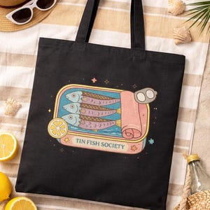 Black sardine tote bag with Tin Fish Society design styled with lemons and beach accessories