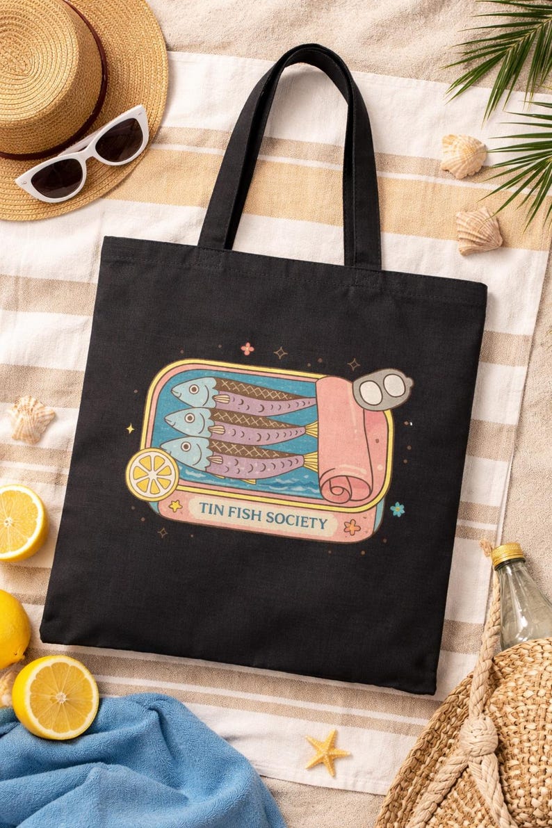 Black sardine tote bag with Tin Fish Society design styled with lemons and beach accessories