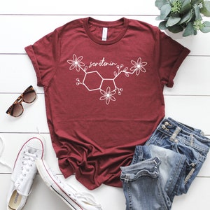 Puede incluir: Una camiseta de color granate con un gr&aacute;fico blanco de la mol&eacute;cula de serotonina con flores. La camiseta est&aacute; colocada sobre una superficie de madera blanca con unas gafas de sol, unas zapatillas blancas y unos vaqueros azules.