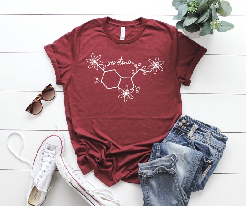 Puede incluir: Una camiseta de color granate con un gr&aacute;fico blanco de la mol&eacute;cula de serotonina con flores. La camiseta est&aacute; colocada sobre una superficie de madera blanca con unas gafas de sol, unas zapatillas blancas y unos vaqueros azules.