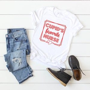 May include: White t-shirt with red text that says "Cupid's Favorite Nurse" with a red heart and arrow below the text.