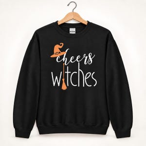 Op de afbeelding: Een zwart sweatshirt met lange mouwen en ronde hals met de witte tekst "cheers witches" op de voorzijde. Het ontwerp bevat een oranje heksenhoed boven het woord cheers en een oranje bezemsteel die de letter i in witches vervangt.