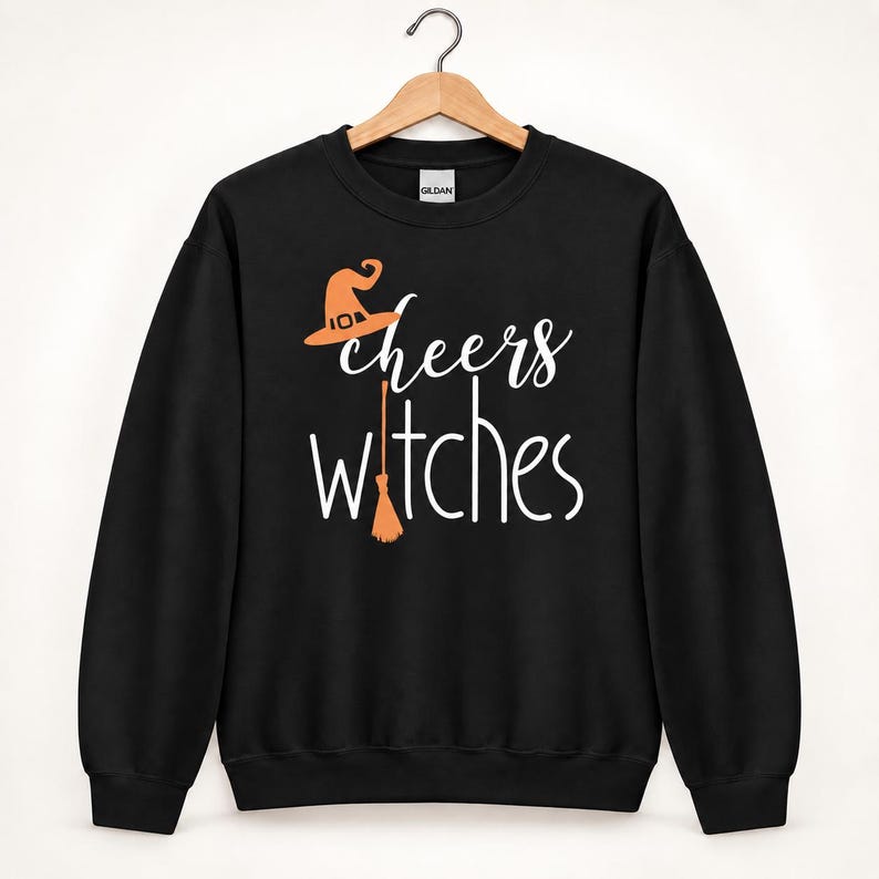 Op de afbeelding: Een zwart sweatshirt met lange mouwen en ronde hals met de witte tekst "cheers witches" op de voorzijde. Het ontwerp bevat een oranje heksenhoed boven het woord cheers en een oranje bezemsteel die de letter i in witches vervangt.