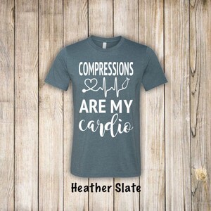 May include: A heather slate blue t-shirt with the text "COMPRESSIONS ARE MY cardio" in white lettering. A heart and EKG line with a stethoscope are included in the design.