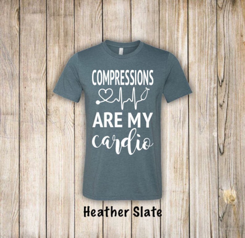 May include: A heather slate blue t-shirt with the text "COMPRESSIONS ARE MY cardio" in white lettering. A heart and EKG line with a stethoscope are included in the design.